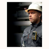 A man in a white hard hat and black jacket stands indoors with a BW Honeywell BW Icon+ multi-gas detector clipped to his attire. The dimly lit background suggests an industrial setting, underscoring the serious nature of his work environment.