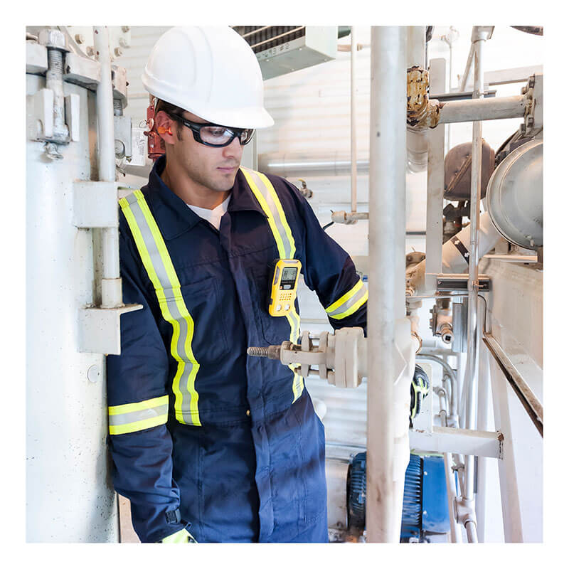 A worker in a navy blue coverall and safety gear inspects equipment, with a BW Technologies by Honeywell BW Clip4 4-Gas Detector BWC4-Y-N clipped to his chest pocket.