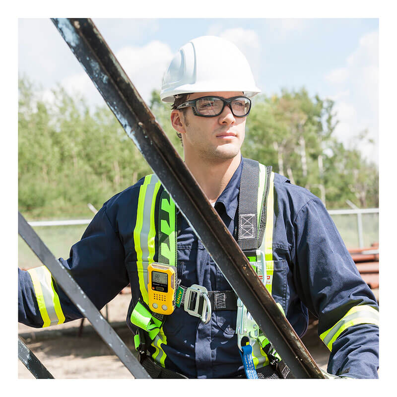 A construction worker in a white hard hat, safety glasses, and reflective vest stands by scaffolding outdoors, equipped with the BW Clip4 4-Gas Detector BWC4-Y-N from BW Technologies by Honeywell, with trees and materials nearby.