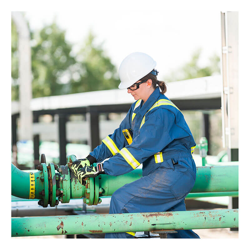 A worker in blue protective gear and a white hard hat, with a BW Technologies by Honeywell BW Clip4 4-Gas Detector BWC4-Y-N clipped on, adjusts an outdoor green industrial pipe. Trees and buildings appear in the background.