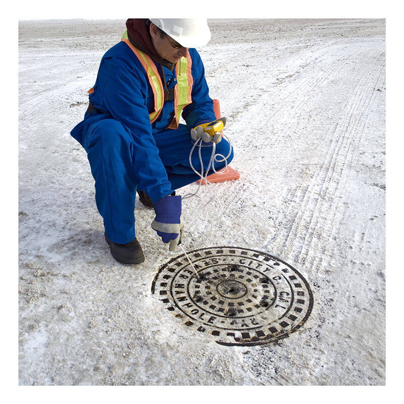 A worker in blue coveralls and a safety vest kneels on snowy ground, using the BW Technologies by Honeywell GasAlert Max XT II Multi-Gas Monitor to inspect a manhole cover labeled "City of Calgary.