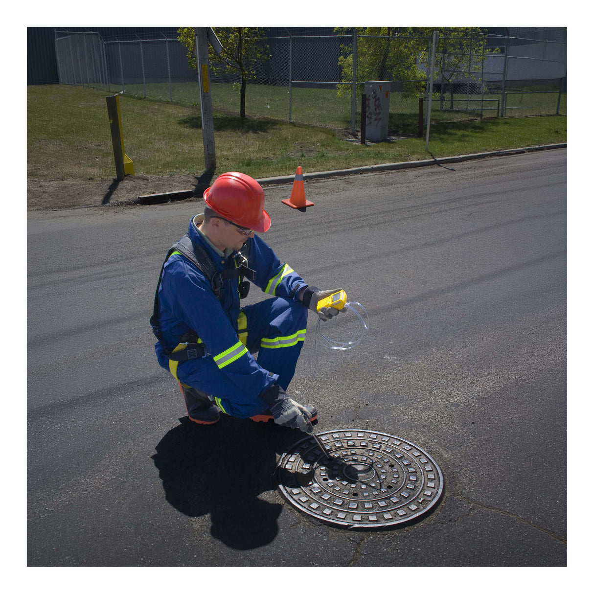 A worker in a helmet and reflective gear kneels by a manhole, inspecting it with a BW Technologies by Honeywell GasAlert Max XT II Confined Space Multi-Gas Monitor. An orange safety cone and green grass are visible in the background.