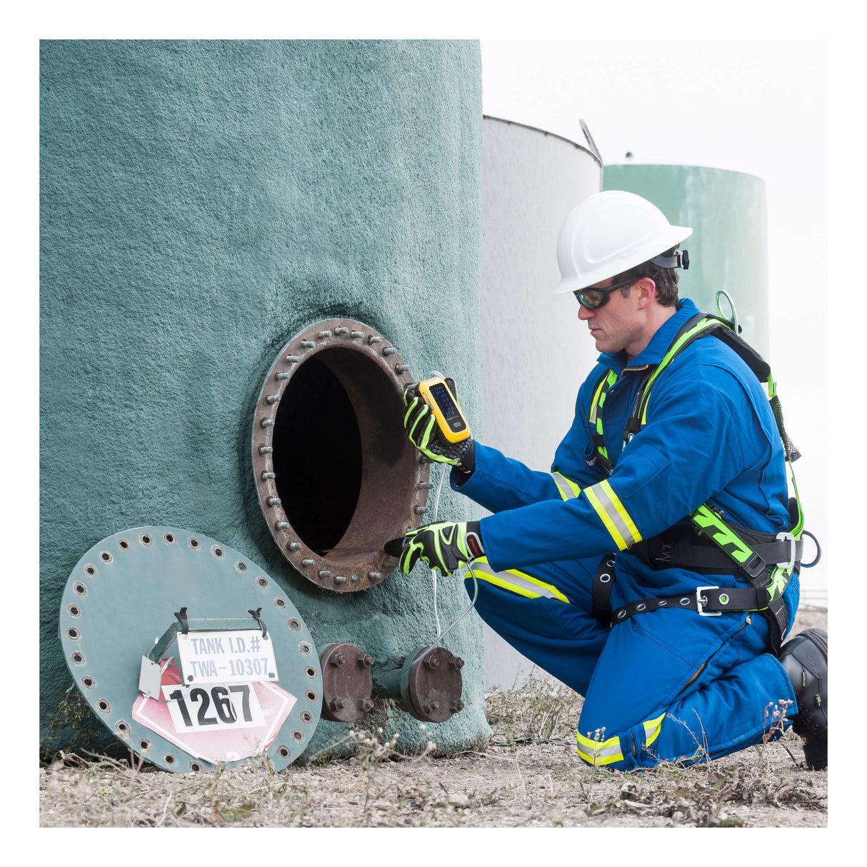 Wearing safety gear and a helmet, a worker kneels by an open tank hatch, using a BW Technologies by Honeywell BW Ultra Five-Gas Portable Gas Detector HU-X1W1H1M1 at the opening. A removed cover and ID sign lie nearby.