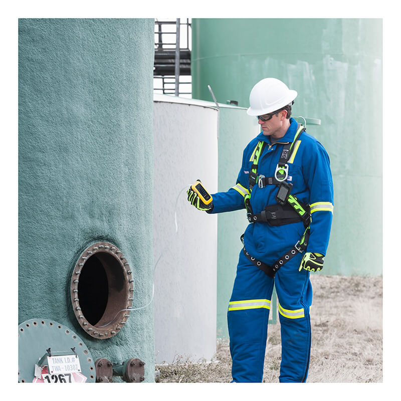 A worker in blue protective gear and a white hard hat uses a BW Technologies by Honeywell BW Ultra Five-Gas Portable Gas Detector (HU-X1W1H1M1) near an open tank hatch outdoors, with safety equipment and warning labels visible.
