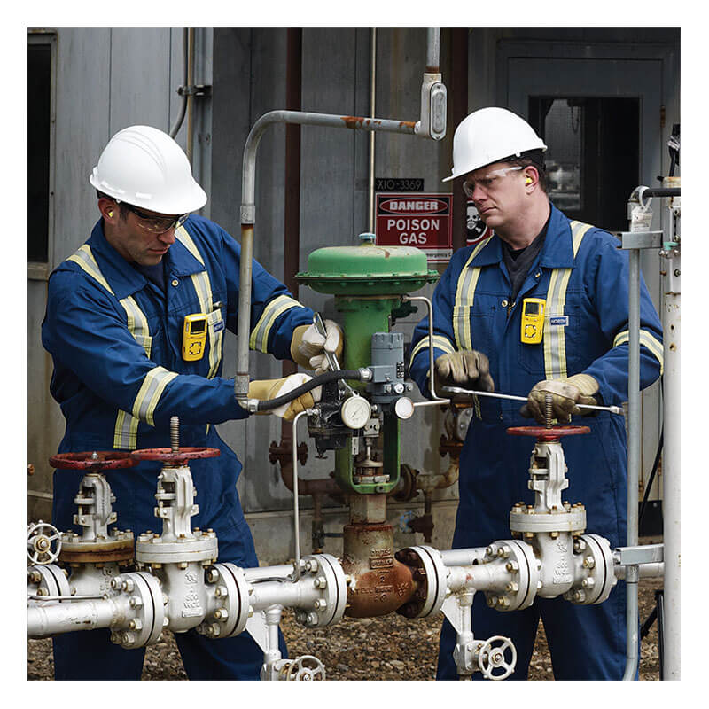 Two workers in blue coveralls and white hard hats inspect outdoor industrial valves with a BW Technologies by Honeywell GasAlert MicroClip XL Multi-Gas Monitor. A “Poison Gas” warning sign is visible in the background.