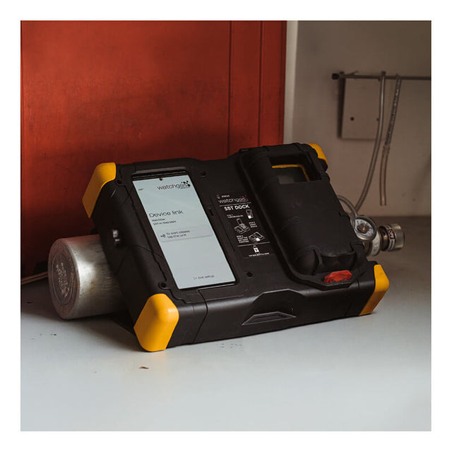 A WatchGas SST Dock Bundle with a black and yellow case rests on its side near a red wall, showing the metal cylinder and regulator—ideal for calibration gas or bump test needs.