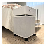 A white Austin Air Systems Bedroom Machine B402 air purifier with a medical-grade HEPA filter sits on a beige carpet next to a bed. Labeled arrows show its size: 23 inches tall and 14.5 inches wide and deep.