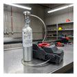 A metal gas cylinder with a pressure gauge is connected via hose to an RKI Instruments SDM-3R Stand Alone Calibration Station (81-SDM3R101) on a workbench in an industrial workshop, with shelves and tools visible in the background.
