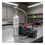 A metal gas cylinder with a pressure gauge is connected via hose to an RKI Instruments SDM-3R Stand Alone Calibration Station (81-SDM3R101) on a workbench in an industrial workshop, with shelves and tools visible in the background.