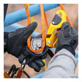 A close-up of gloved hands using the Petzl RIG Compact Descender D021_ _00 to attach an orange rope to its AUTO-LOCK system with yellow and black parts, set against a blue metal structure in the background.