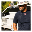 A man wearing a hard hat, sunglasses, and a navy blue NSA DRIFIRE Short Sleeve FR Base Layer T-Shirt (DF2-CM-446TS) stands outdoors near a white utility vehicle, looking to the side.