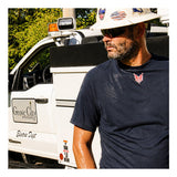 A man wearing a hard hat, sunglasses, and a navy blue NSA DRIFIRE Short Sleeve FR Base Layer T-Shirt (DF2-CM-446TS) stands outdoors near a white utility vehicle, looking to the side.