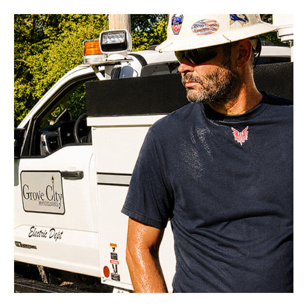 A man wearing a hard hat, sunglasses, and a navy blue NSA DRIFIRE Short Sleeve FR Base Layer T-Shirt (DF2-CM-446TS) stands outdoors near a white utility vehicle, looking to the side.
