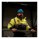 A bearded person in a blue hard hat and yellow safety jacket stands indoors, holding a railing and looking ahead in an industrial setting, equipped with the WatchGas SST5 Diffusion 5-Gas Detector LPC/O2/CO/H2S/Exotic by WatchGas to ensure safety.