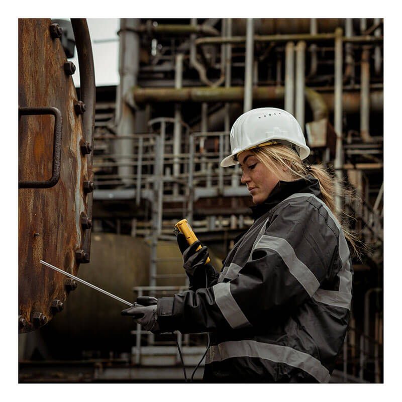 Wearing safety gear, including a white helmet and gloves, a woman uses the WatchGas SST4-PUMP 4-Gas Detector to inspect industrial equipment at a factory with metal structures and pipes in the background.