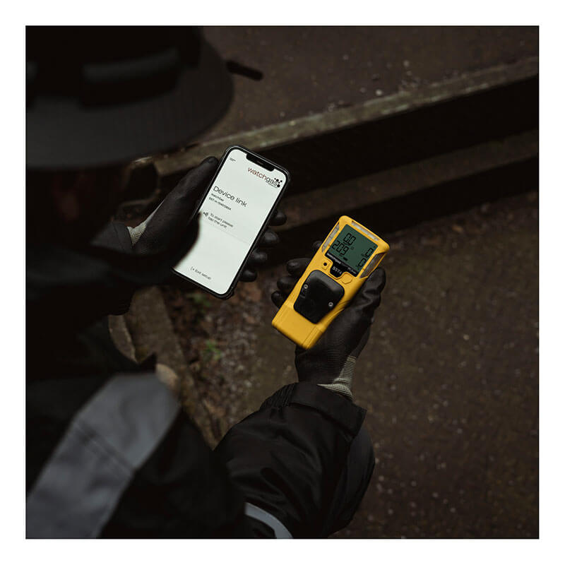 A person wearing gloves stands outdoors near concrete steps, holding a smartphone showing a service report in one hand and a yellow WatchGas SST4-PUMP 4-Gas Detector by WatchGas with a digital screen in the other.