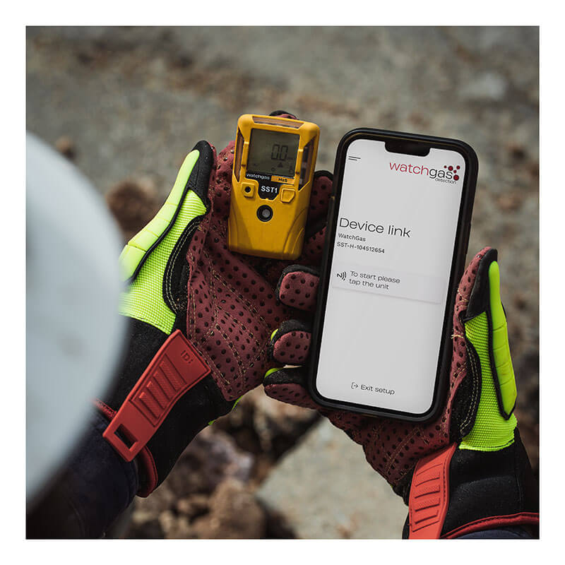 A person wearing protective gloves holds a WatchGas SST1 Serviceable CH4S Gas Detector SST1-MM in one hand and a smartphone with a gas monitoring app in the other, with rocky ground visible in the background.