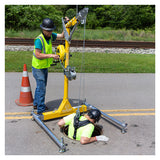 Two workers in safety gear use the French Creek Production Portable Davit System Winch Mounting Bracket PD4 near a manhole; one operates the winch while the other enters a confined space. A traffic cone and railroad tracks can be seen in the background.