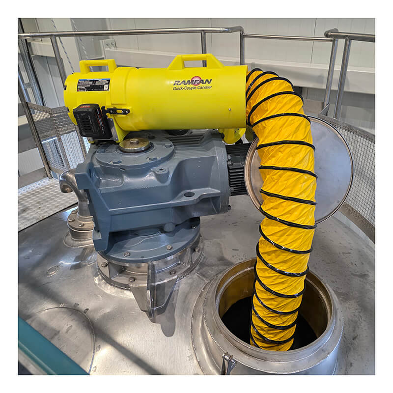 A Euramco RAMFAN UB20 18VDC and Quick-Couple Canister, an essential ventilation component from Euramco, is connected to industrial equipment with a flexible yellow hose, leading into an open metal tank in a facility.