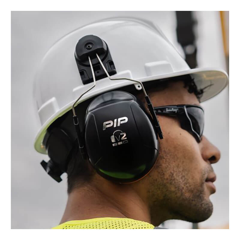 A person wearing a white hard hat, PIP V2 Full Brim Mounted Ear Muff 263-V2FB (NRR 25), black safety glasses, and a yellow safety vest—showcasing key PIP safety equipment at a worksite.