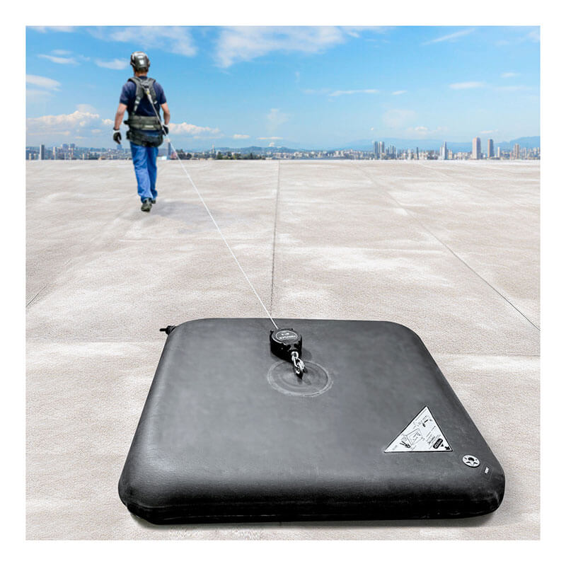 A worker in safety gear walks on a flat rooftop, attached to a retractable safety line connected to the Frontline EcoAnchor Counterweight Fall Arrest Anchor System by Frontline Fall Protection. City buildings are visible in the distance.
