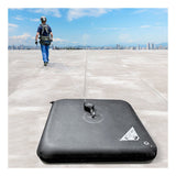 A worker in safety gear walks on a flat rooftop, attached to a retractable safety line connected to the Frontline EcoAnchor Counterweight Fall Arrest Anchor System by Frontline Fall Protection. City buildings are visible in the distance.