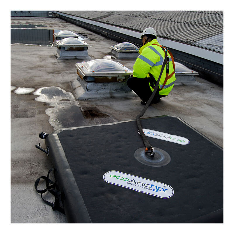 A worker in a high-visibility jacket and safety harness is crouched on a flat rooftop near skylights, secured by a line to the Frontline EcoAnchor Counterweight Fall Arrest Anchor System from Frontline Fall Protection.