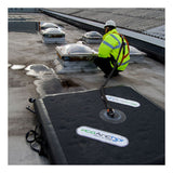A worker in a high-visibility jacket and safety harness is crouched on a flat rooftop near skylights, secured by a line to the Frontline EcoAnchor Counterweight Fall Arrest Anchor System from Frontline Fall Protection.