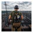 A construction worker in a hard hat uses the French Creek 8’ Class 2 Personal SRL XR_-8LE by French Creek Production while standing on a high platform, overseeing an industrial site with steel structures and cranes beneath a cloudy sky.