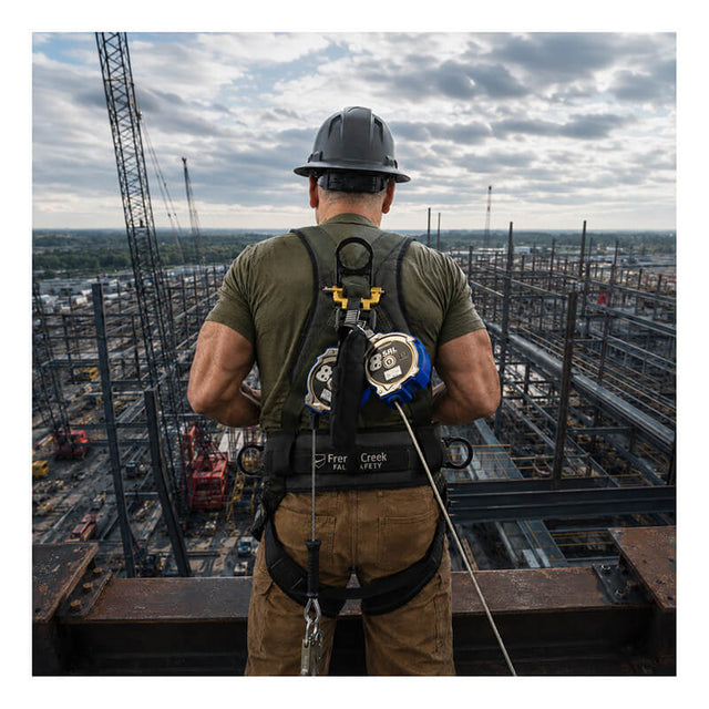A construction worker in a hard hat uses the French Creek 8’ Class 2 Personal SRL XR_-8LE by French Creek Production while standing on a high platform, overseeing an industrial site with steel structures and cranes beneath a cloudy sky.