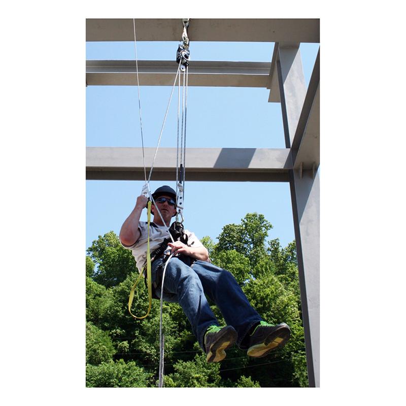 Wearing safety gear and sunglasses, a person is suspended in mid-air using ropes and pulleys from a French Creek Production System 99 Self-Raising/Lowering System 99B. Positioned beneath a metal structure with trees and a clear blue sky in the background, they showcase an efficient lifting and lowering system.