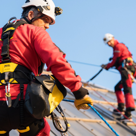 Two workers in red safety vests and helmets on a roof, one using a tool.