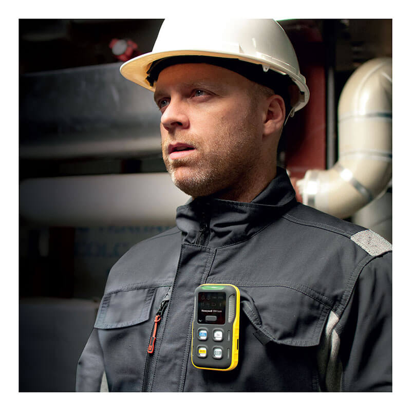 A construction worker wearing a white hard hat and dark jacket stands indoors with a yellow BW Flex 4 %LEL, O2, H2S, CO gas monitor from BW Technologies by Honeywell clipped to his chest, focused amid visible industrial pipes.