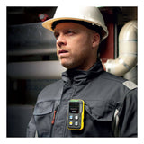 A construction worker wearing a white hard hat and dark jacket stands indoors with a yellow BW Flex 4 %LEL, O2, H2S, CO gas monitor from BW Technologies by Honeywell clipped to his chest, focused amid visible industrial pipes.