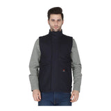 A man with short brown hair wears a ForgeFR Forge FR Canvas Duck Vest MFRVST-001 (NFPA2112-compliant, black) over a light gray patterned shirt and blue jeans, standing and facing forward against a plain white background.