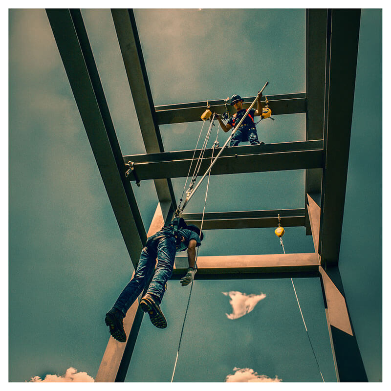 A construction worker uses the French Creek Rescue Pole RPCC-12 by French Creek Production while suspended in fall protection gear, secured with a carabiner, against a blue sky on a steel framework.