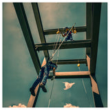 A construction worker uses the French Creek Rescue Pole RPCC-12 by French Creek Production while suspended in fall protection gear, secured with a carabiner, against a blue sky on a steel framework.