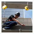A construction worker in a hard hat, safety glasses, harness, and jeans is crouched on a steel beam high up, secured by a French Creek Production 50ft Galvanized SRL RL50GZ. A city building rises in the background.