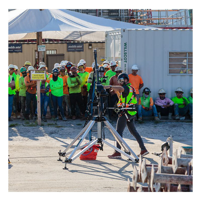 A worker in a safety vest operates equipment near a Frontline Fall Protection Patriot CS Quadpod with 3-Way Retrieval System at a construction site, while others in hard hats and high-visibility clothing observe a nearby "Caution" sign.