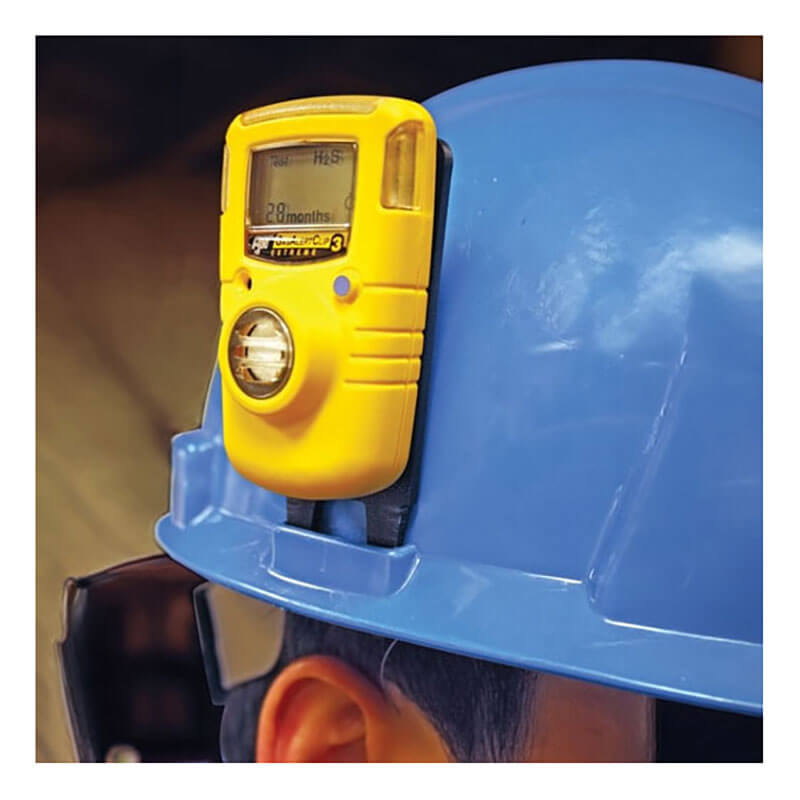 A hard hat, in blue, is worn by a worker with a yellow gas detector securely attached using the BW Hard Hat Clip GA-HC-1 from PK Safety Supply. The device's screen shows "28 months" and an "H2S" level for efficient gas detection in the field.