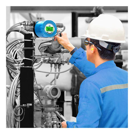 A worker in a blue uniform and white helmet operates the GfG CC33 CH4 Fixed Transmitter Aluminum 3300-41-00, which is equipped with numerous knobs and cables. The digital display on this GfG equipment reads "25.0." The worker, wearing ear protection and glasses, diligently ensures safety in an area sensitive to explosion hazards.