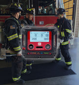 Three firefighters in full gear stand near a fire truck inside a station. An overlay shows the RKI Instruments GX-3R Pro 5 Gas Confined Space Monitor with Wireless Communication displaying air quality readings.