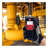 A gloved hand holds an RKI Instruments GX-Force Sample Draw 4 Gas Monitor (72-FA-A-A10) in front of yellow industrial pipes and equipment. The display shows precise gas measurements, indicating use inside a facility or plant.