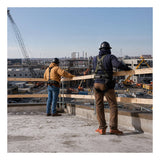 Two construction workers equipped with safety harnesses and hard hats stand on a rooftop, surveying an active construction site filled with cranes, scaffolding, and building materials beneath a clear sky. The French Creek Production Portable Guardrail System GR200 is installed along the roof edge, underscoring their dedication to fall prevention on the jobsite.