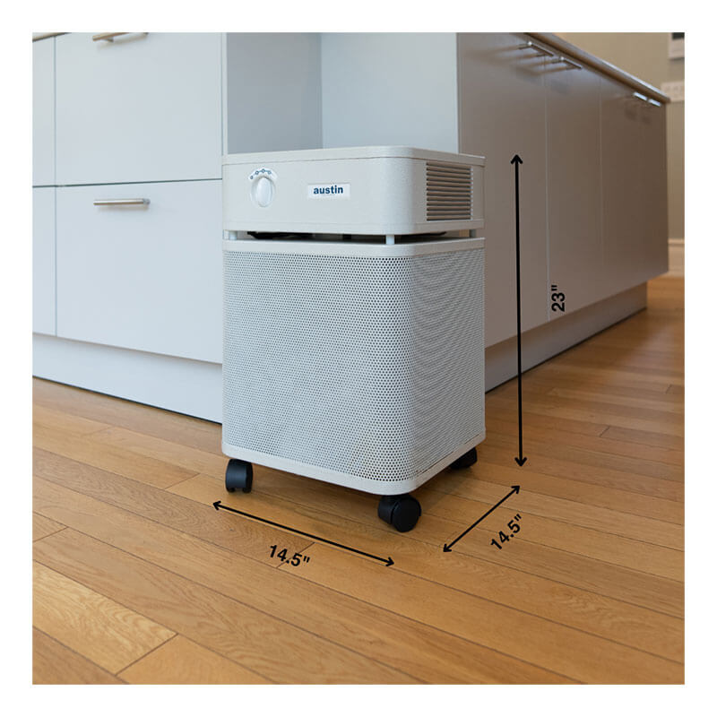 A white, box-shaped Austin Air Systems HealthMate Machine B400 air purifier with wheels sits on a hardwood floor near kitchen cabinets. Black arrows show its dimensions: 23" tall, 14.5" deep, 14.5" wide; features medical grade HEPA filter.
