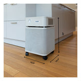 A white, box-shaped Austin Air Systems HealthMate Machine B400 air purifier with wheels sits on a hardwood floor near kitchen cabinets. Black arrows show its dimensions: 23" tall, 14.5" deep, 14.5" wide; features medical grade HEPA filter.