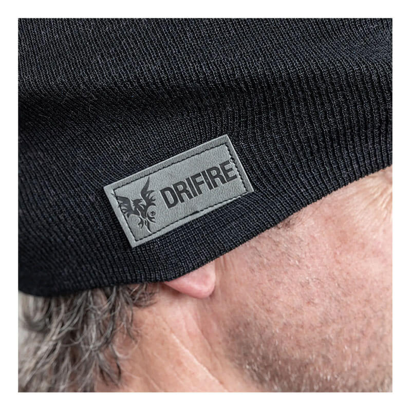Close-up of a person wearing the NSA DRIFIRE FR Winter Ear Band HNCEB, a black flame-resistant knit beanie with a gray "DRIFIRE" patch featuring a flame logo. The person's ear and some gray hair are visible.
