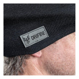 Close-up of a person wearing the NSA DRIFIRE FR Winter Ear Band HNCEB, a black flame-resistant knit beanie with a gray "DRIFIRE" patch featuring a flame logo. The person's ear and some gray hair are visible.