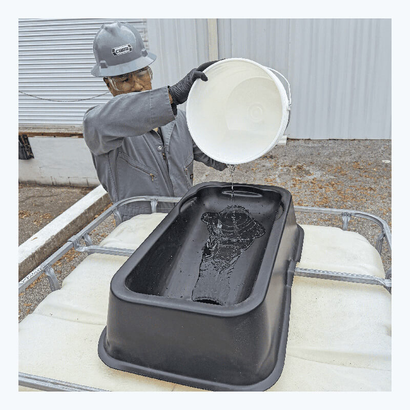 Wearing safety gear, a person uses the UltraTech IBC Mini Funnel 0649 to pour liquid from a white bucket into a black plastic container on top of an outdoor white storage tank.