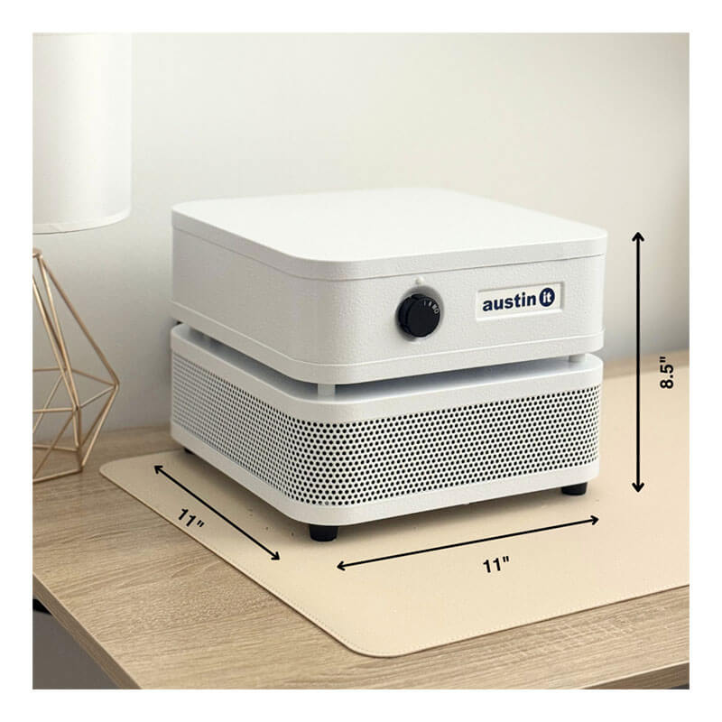 The white Austin Air "it" Machine C100 by Austin Air Systems sits on a desk mat. Measuring 8.5" tall by 11" wide and deep, it features a control knob and logo, designed to remove airborne particles and VOCs.